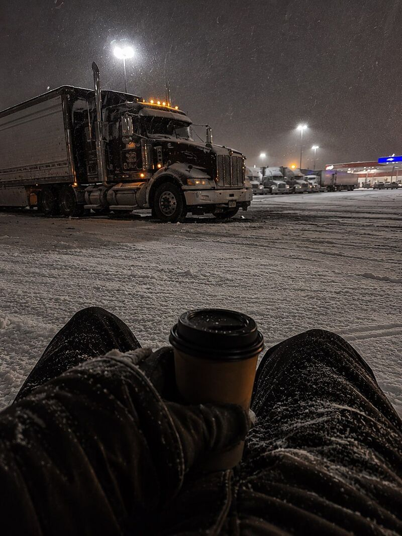 A driver's view from the curb of a snowy midnight truck stop, holding a paper coffee cup as a Peterbilt idles under the lot lights.